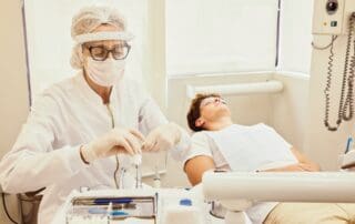 Dentist in surgical attire preparing dental instruments for a patient lying comfortably on the bed.