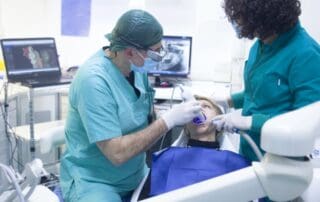 Senior dentist in surgical uniform, assisted by his assistant, placing a medical instrument into the patient's mouth during dental care.