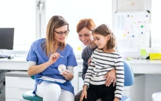 Female dentist showing dental instruments to a child while sitting on the mother's lap.